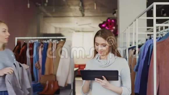 年轻的企业主站在她的服装店里使用平板电脑她的助手带着衣服来视频的预览图