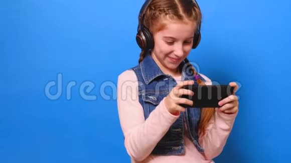 戴着耳机的小女孩在智能手机上玩电子游戏微笑着视频的预览图