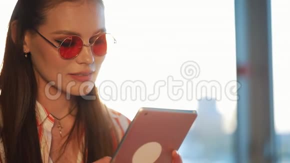 年轻迷人的女士戴着红色太阳镜在咖啡馆里使用带有触摸屏的平板电脑机场或机场的美女视频的预览图