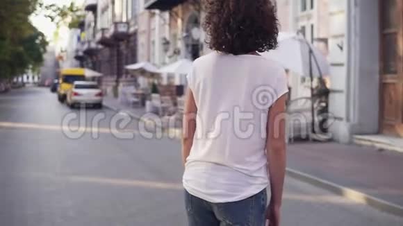 城市街上一个穿着白色t恤蓝色牛仔裤和白色运动鞋的女人的后景你的腿视频的预览图