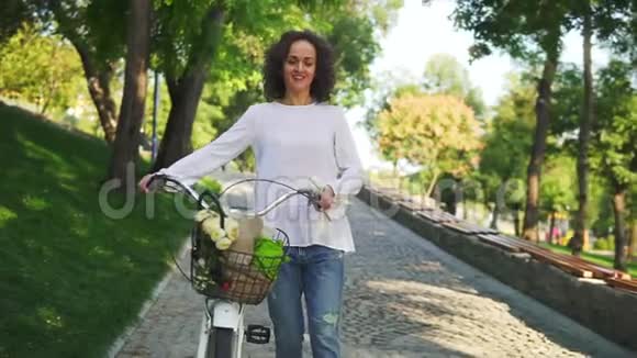 穿着白色T恤和蓝色牛仔裤的迷人女人微笑着手里拿着她的城市自行车带着鲜花的车把视频的预览图