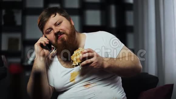 肮脏的胖胡子男人在智能手机上聊天吃汉堡坐在沙发上喝啤酒概念视频的预览图