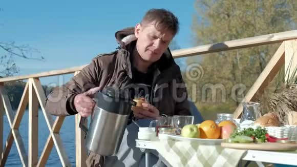 成熟的人穿着温暖的衣服坐在桌子旁吃东西从热水瓶里倒茶在河岸上野餐视频的预览图