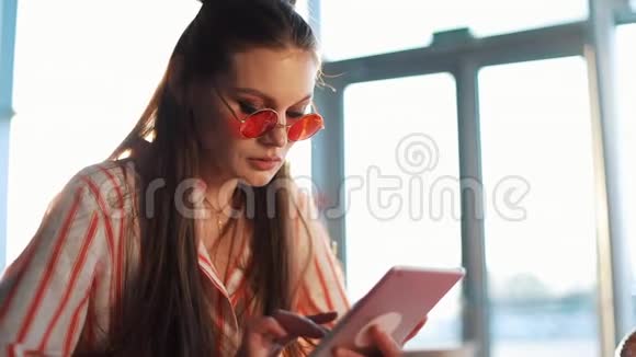 年轻迷人的女士戴着红色太阳镜在咖啡馆里使用带有触摸屏的平板电脑机场或机场的美女视频的预览图