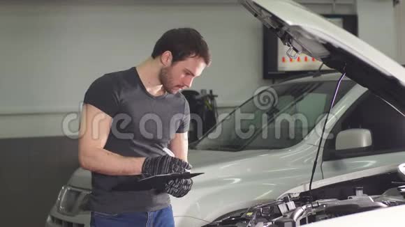 汽车机械师站在汽车附近的一家汽车修理店里开着引擎盖做着笔记视频的预览图