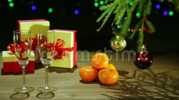 圣诞树旁边的香槟酒杯和带有灯光的黑色背景的木桌上的礼物视频的预览图