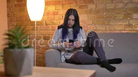 年轻漂亮的白种人潮女在室内坐在沙发上用手机特写镜头视频的预览图