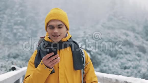 一位年轻的游客站在雪山上的一座桥上手里拿着一部智能手机交流社会视频的预览图