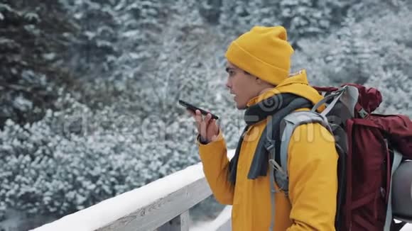 一位年轻的游客站在雪山上的一座桥上手里拿着一部智能手机徒步旅行者和视频的预览图