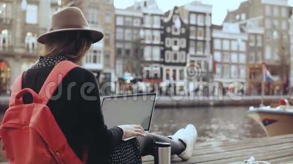 4K年轻美女博客使用笔记本电脑阿姆斯特丹自由戴着时髦帽子的红色背包的女孩在河堤上视频的预览图