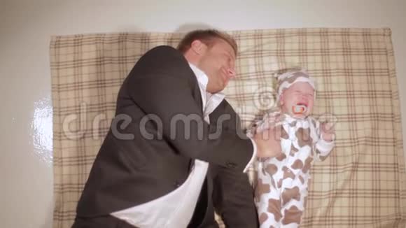 穿着西装的成熟白种人躺在棕色毯子上玩着尖叫的婴儿视频的预览图