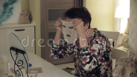 一位老妇人看着镜子里的皱纹她打开一个化妆织物面具把它放在她的脸上面部护理视频的预览图