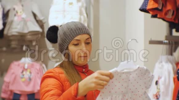 宝贝服装店里漂亮的女人戴着灰色针织帽子和红色夹克的白种人女性手里拿着婴儿装视频的预览图