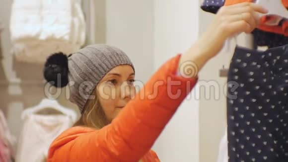 宝贝服装店里漂亮的女人穿着灰色针织帽子和红色夹克的白人女性看着婴儿裤视频的预览图