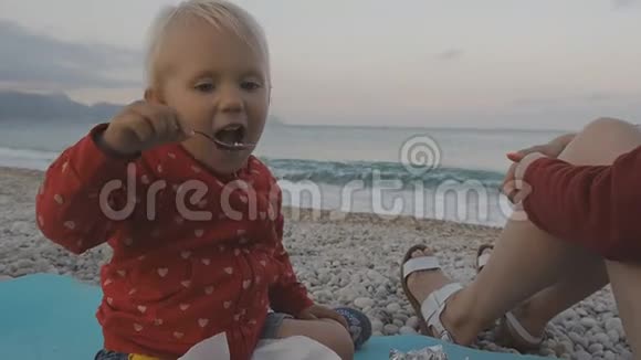 两岁的婴儿用勺子吃食物在海滩上咀嚼孩子和妈妈在日落海滩上视频的预览图