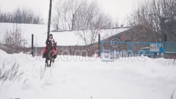 穿着红色连衣裙的年轻黑发女郎在冬天骑着马在白雪覆盖的田野里快速慢跑视频的预览图