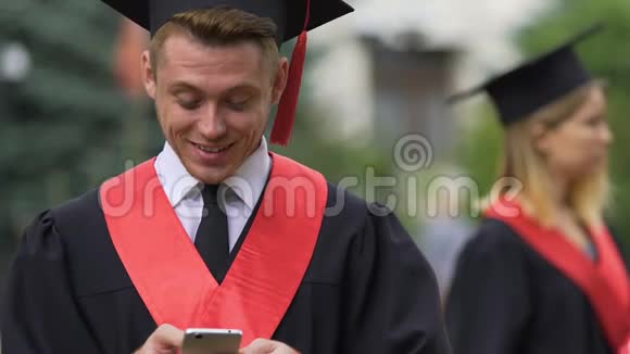 开朗的男性毕业生穿着学术服装在智能手机上聊天笑着视频的预览图