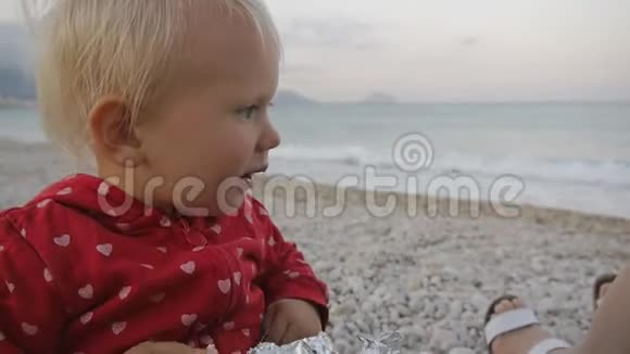 两岁婴儿在海边背景上咀嚼食物的肖像孩子和妈妈在日落海滩上视频的预览图