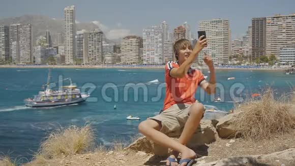 白种人少年在智能手机上打视频电话展示海上海岸和摩天大楼天际线背景视频的预览图