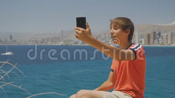 英俊的微笑少年在海上海岸和摩天大楼天际线背景下制作智能手机视频通话的肖像视频的预览图