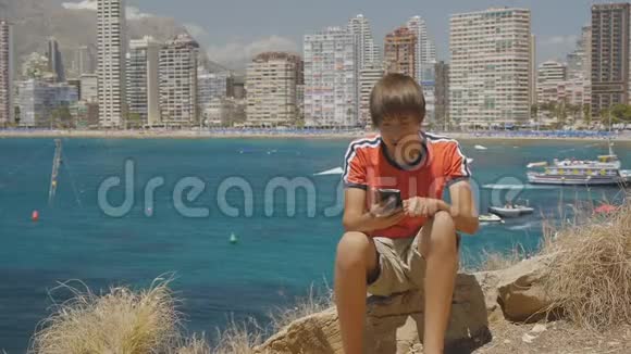 15岁的男孩坐在海边的石头上在摩天大楼的天际线背景下用智能手机输入信息视频的预览图