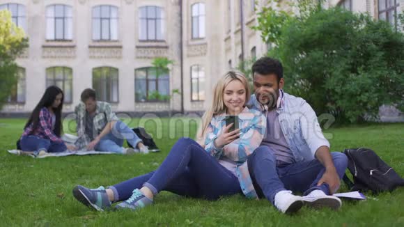 可爱的几个学生坐在草坪上看智能手机上的视频视频的预览图