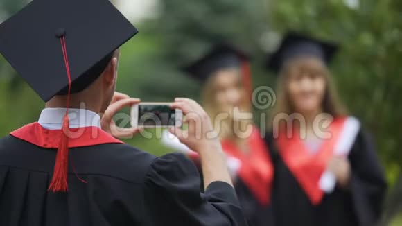 年轻毕业生披着斗篷戴着帽子用手机拍照友谊视频的预览图