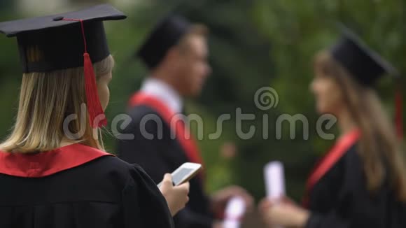 穿着学术服装和戴毕业帽的金发女孩在智能手机上上网视频的预览图