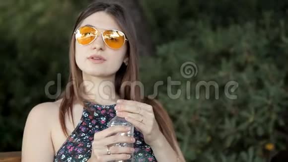一个年轻的女孩坐在公园的长凳上喝水视频的预览图