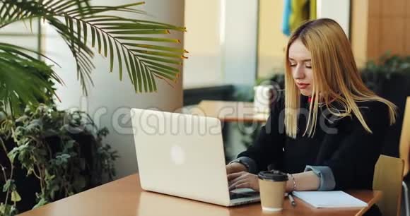 年轻的金发女孩在咖啡馆的桌子上和一台笔记本电脑一起工作自由职业者正在找工作打印一些东西视频的预览图