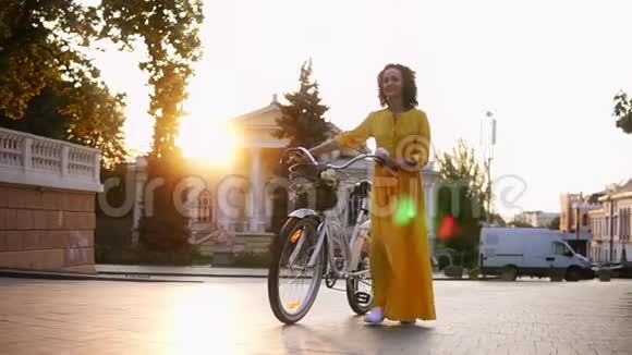 美丽的年轻女子穿着一件黄色的长裙在黎明时分手里拿着她的自行车带着鲜花的车把视频的预览图