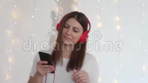 美丽的女孩听音乐耳机红色坐在床上放松使用手机特写视频的预览图