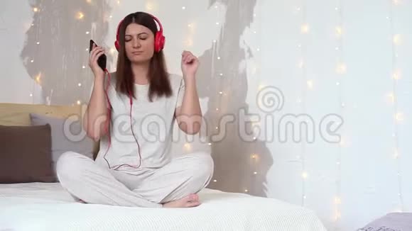 一个穿着睡衣的漂亮女孩放松下来一边在耳机上听音乐一边心情很好地坐在床上视频的预览图