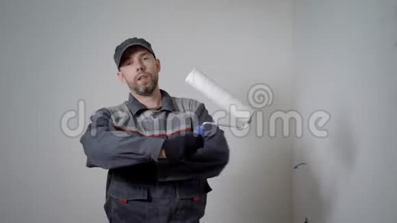 开朗的修理工站在公寓里拿着油漆滚筒双手交叉在白色空墙的背景上视频的预览图