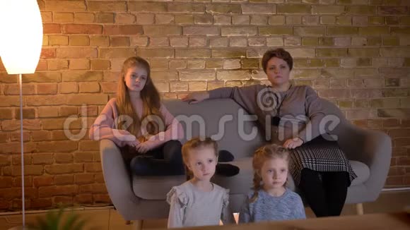 白种人母亲的家庭肖像三个女儿坐在沙发上在舒适的家庭氛围中看电影视频的预览图