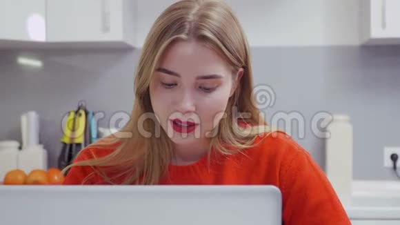 穿红色毛衣的年轻女孩在厨房用笔记本电脑视频的预览图