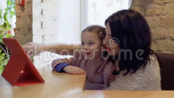 妈妈和小女儿坐在咖啡馆里看着平板电脑上的卡通片视频的预览图
