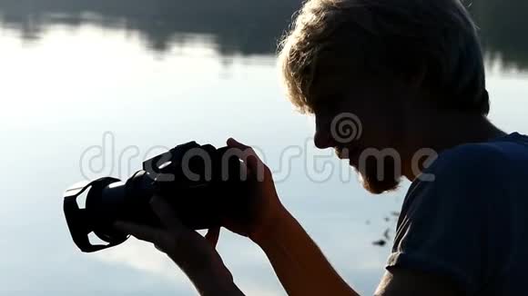 金发男人坐在森林湖边夏天看着他的相机视频的预览图