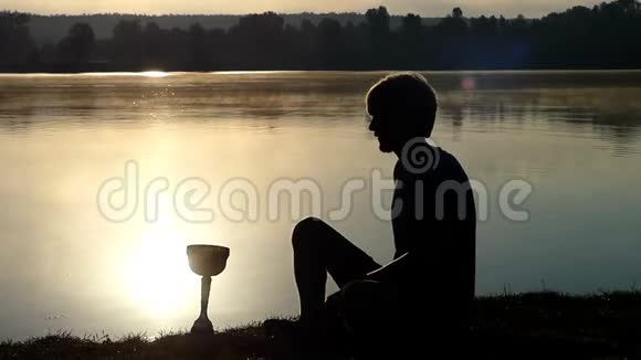 金发碧眼的人坐在湖岸上看着一只盛满水的冠军碗视频的预览图