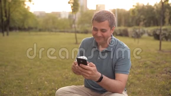 微笑的男人坐在公园的草坪上喝着咖啡打着电话和一个朋友聊天视频的预览图