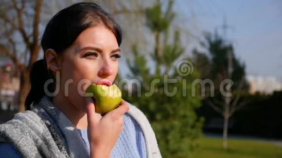 在阳光明媚的日子里有着美丽的眼睛和红唇的迷人年轻黑发女人吃着绿苹果视频的预览图