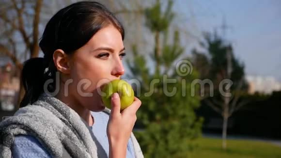 在阳光明媚的日子里有着美丽的眼睛和红唇的迷人年轻黑发女人吃着绿苹果视频的预览图