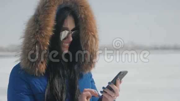 戴着头巾戴着墨镜戴着智能手机的年轻美丽黑发女人的肖像在冬天的雪地背景下使用智能手机视频的预览图