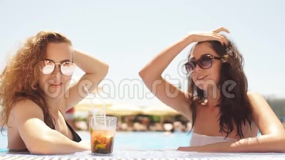 两个年轻可爱的女朋友戴着太阳镜穿着泳衣在游泳池里喝着鸡尾酒杯户外肖像画视频的预览图