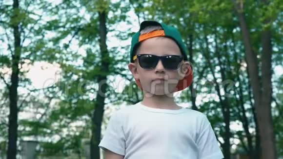 一个戴着太阳镜的酷男孩展示了一个班他的手指站在一个夏天的公园里背景是绿树视频的预览图