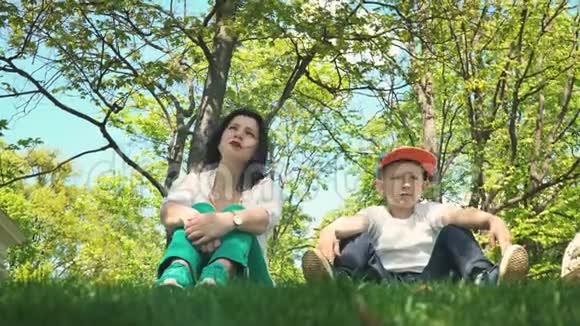 妈妈和儿子正看着远处一边聊天一边坐在公园的绿草上一家人度假视频的预览图