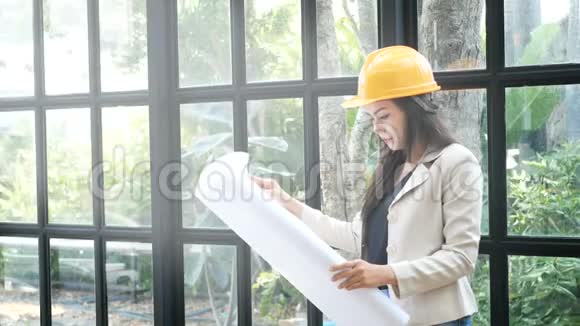 年轻的亚洲女建筑工程师戴着黄色的安全帽手里拿着建筑师的蓝图视频的预览图