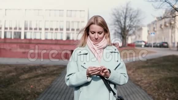 快乐的白人30多岁的女人在智能手机上打字在城里散步时发短信的金发美女慢动作视频的预览图