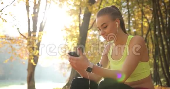 公园里的女跑步者穿着慢跑服的可爱的年轻女子在跑步后休息听音乐视频的预览图