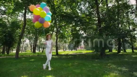 快乐的小女孩在阳光明媚的绿色公园里摆姿势手里拿着五颜六色的气球视频的预览图